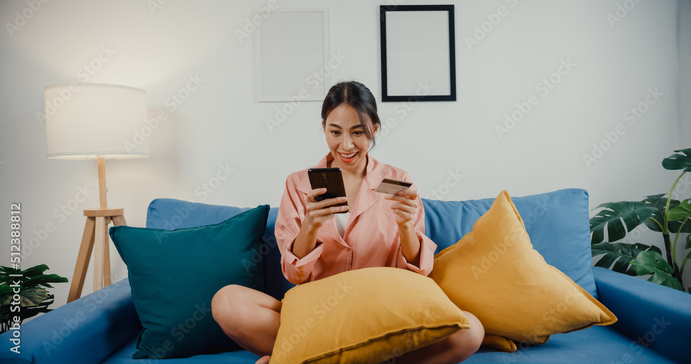 Obraz premium Happy young asia lady use cellphone order online shopping product and paying bill with credit card sit on couch in living room at house. Quarantine activity, Fun activity for coronavirus prevention.