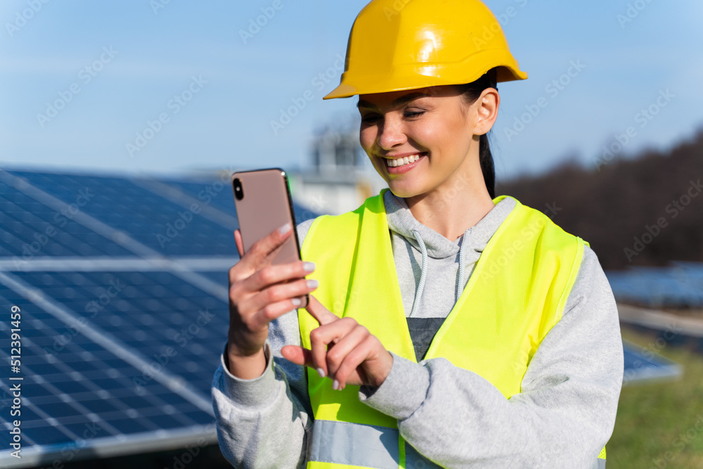 Portrait view of the woman in hard hat using smartphone while relaxing during her break on the background of solar panels Portrait view of the woman in hard hat using smartphone while relaxing during her break on the background of solar panels