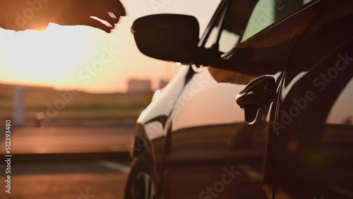 Young attractive woman with paper bags opens the door of red car and gets behind the wheel in parking lot at sunset. Girl in light relaxed clothes, happy after shopping, slow motion. Cinematic footage