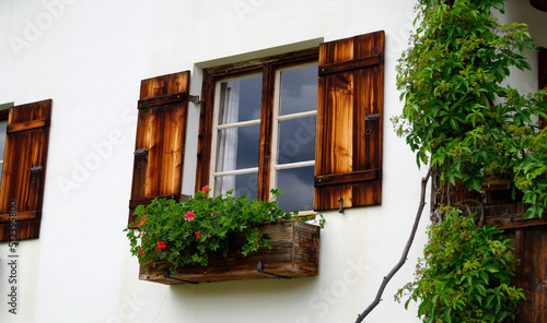 a rustic window with cute white curtains, wooden shutters and red geraniums on the window ledge in the Bavarian alpine countryside Schwangau in the German Alps, Bavaria, Germany