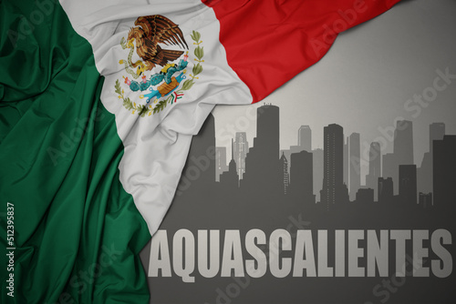 abstract silhouette of the city with text Aguascalientes near waving colorful national flag of mexico on a gray background.