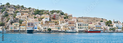 Fototapeta Naklejka Na Ścianę i Meble -  Multi-colored houses scattered on hills of Symi island view from water. Coastal town on turquoise sea shore in Greece. Tourist liners moor by seafront
