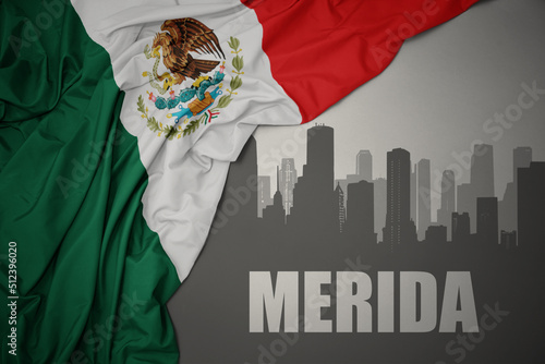 abstract silhouette of the city with text Merida near waving colorful national flag of mexico on a gray background.