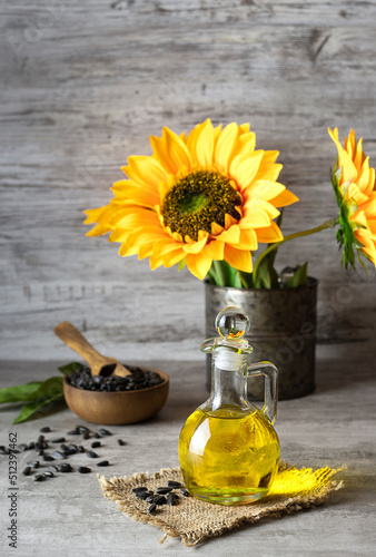Transparent jug with sunflower oil on a canvas napkin. A wooden bowl with seeds in the background. Sunflowers in a tin can on the table. Light wooden background