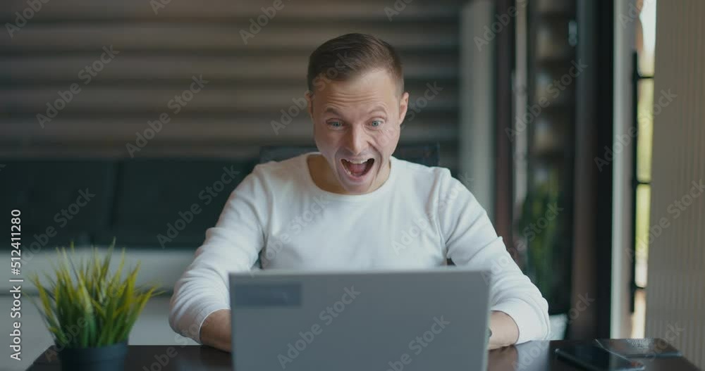 Happy young man sits in room behind laptop and rejoices the good news, says wow. Actively gestures with hands Happy young man sits in room behind laptop and rejoices the good news, says wow. Actively gestures with hands