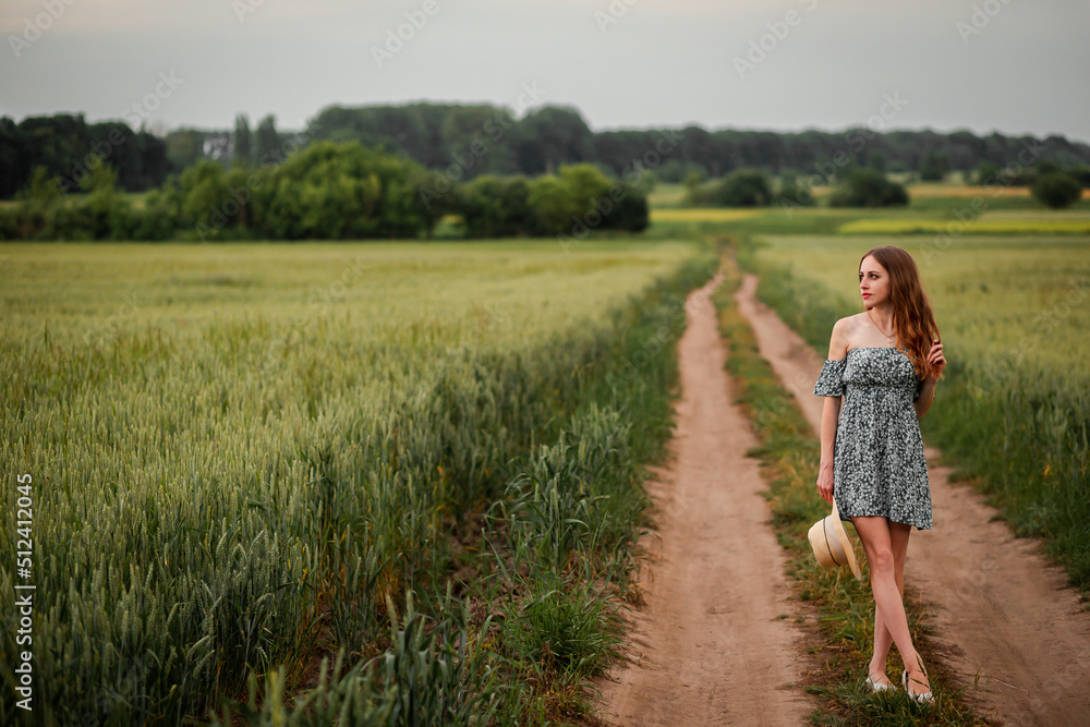 Vinnytsia, Ukraine. May 25, 2022. A girl in a green dress and holding a hat and standing in a wheat field and looking at the sky