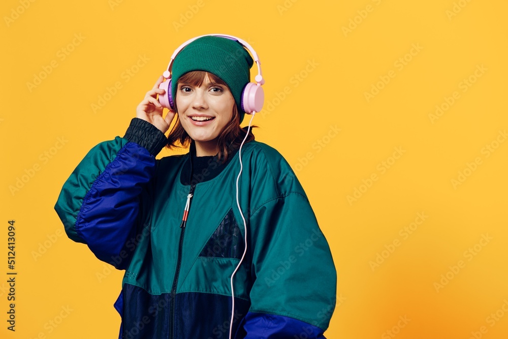 a happy, joyful woman in a green jacket and hat enjoys music standing in pink headphones on a yellow background, holding them with her hands and looking at the camera. Horizontal photo a happy, joyful woman in a green jacket and hat enjoys music standing in pink headphones on a yellow background, holding them with her hands and looking at the camera. Horizontal photo