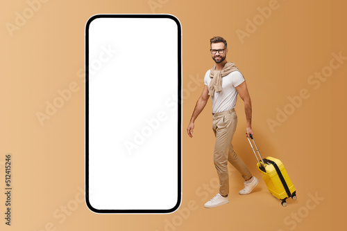 Mobile app for travelers. Handsome man with yellow luggage suitcase walking to huge smartphone with empty display, copy space, mockup