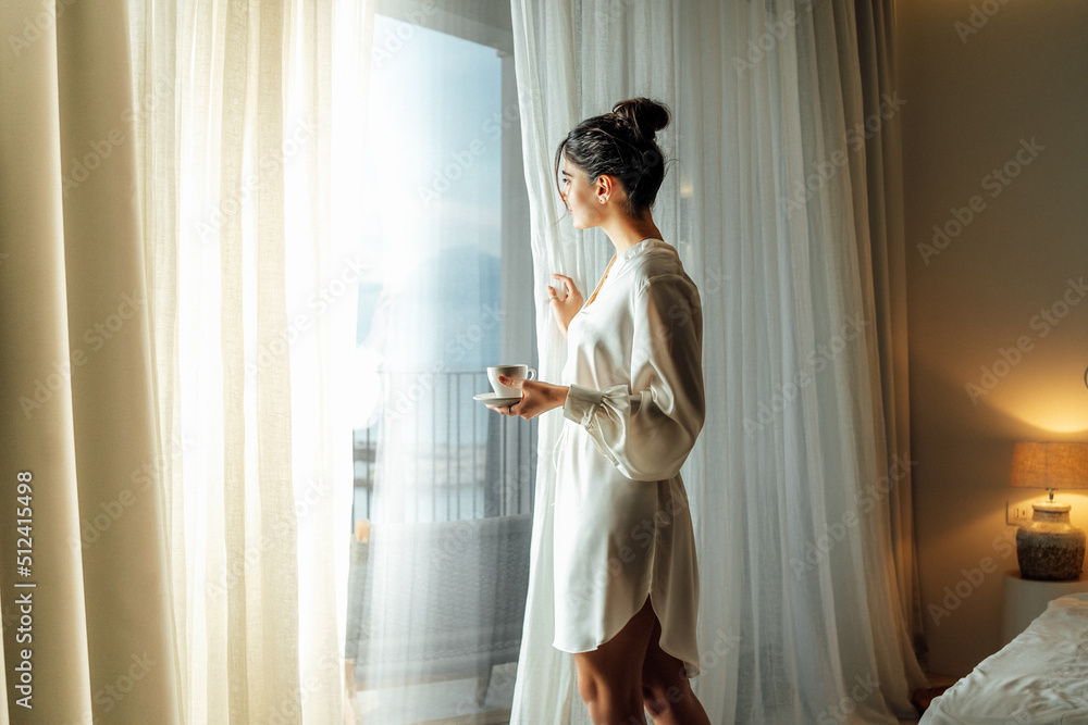 Young woman wearing white bathrobe opening curtains in luxury hotel room and enjoying sun in the morning while having her cup of coffee - obrazy, fototapety, plakaty 