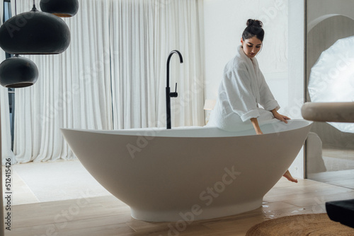 Full length view of the romantic girl preparing to taking bathtub at her cozy apartments. Brunette woman wearing bathrobe sitting at the tub and touching water with her hand
