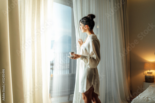 Young woman wearing white bathrobe opening curtains in luxury hotel room and enjoying sun in the morning while having her cup of coffee
