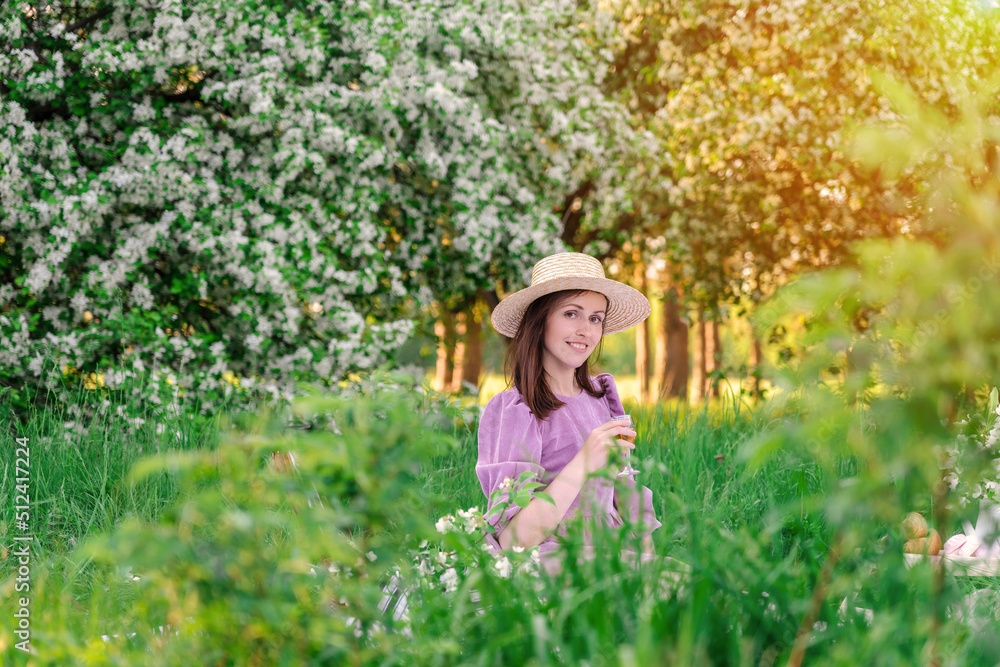 A brunette woman in a purple dress and a straw hat holds a glass of white wine and poses at a picnic in an apple orchard at sunset A brunette woman in a purple dress and a straw hat holds a glass of white wine and poses at a picnic in an apple orchard at sunset