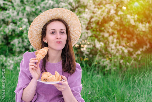 A brunette woman in a purple dress and a straw hat eats fresh croissants at a picnic