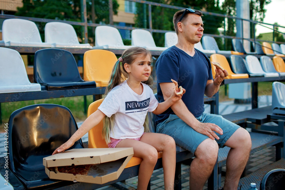 Father with daughter, soccer fans, watching football match, eating pizza and cheering for local team at stadium, real emotions, sports event and fan supporting, outdoor lifestyle, street food