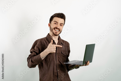 Cheerful caucasian man wearing brown shirt posing isolated over white background smiling and pointing with index finger to the laptop pc computer.
