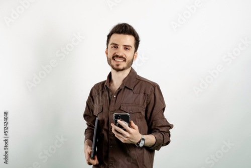 Portrait of young handsome man wearing brown shirt posing isolated over white background holding in hands carrying laptop and phone posing. Looking at the camera with big smiles.