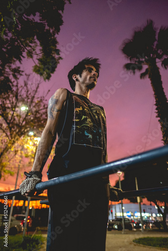 Fit and handsome athletic latin caucasian guy with tattoos and workout gloves doing a calisthenic exercise routine (performs dips on parallel bars) in an street workout park with colorful sunset sky