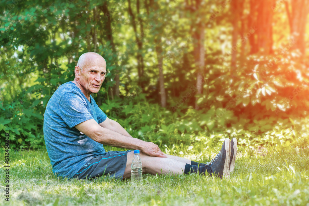 Old tired but happy man seated on park lawn surface is exercising lean and stretching and looking to camera at sunny early morning Old tired but happy man seated on park lawn surface is exercising lean and stretching and looking to camera at sunny early morning