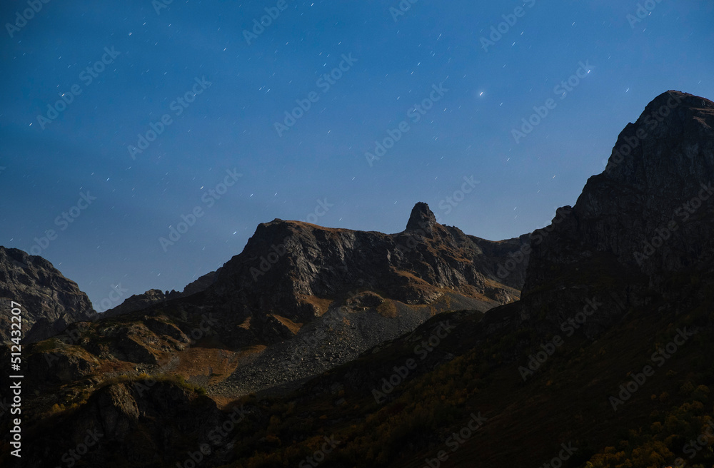 Starry night sky and peaks of rocky mountains in moon light in Caucasus national park. Caucasus nature reserve. Alpine landscape.