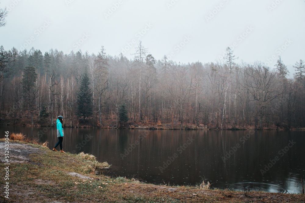 Hiker in a grey and blue jacket at the edge of a pond in rainy weather, enjoying the misty sunset light. Biodiversity of Czech nature. Young pretty brunette discovering new landscapes