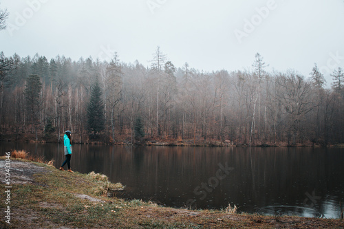 Hiker in a grey and blue jacket at the edge of a pond in rainy weather, enjoying the misty sunset light. Biodiversity of Czech nature. Young pretty brunette discovering new landscapes © Fauren