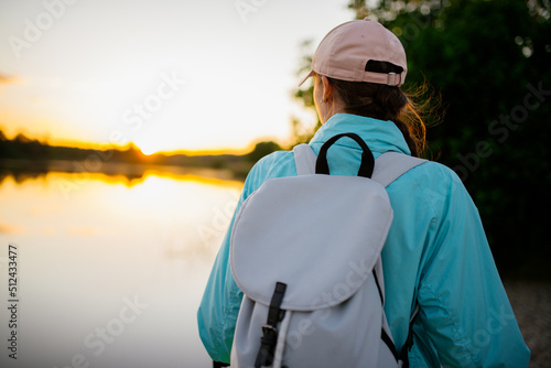 Traveling woman with backpack and hat looking at river at sunset. View from front tourist traveler bag. © prochkailo