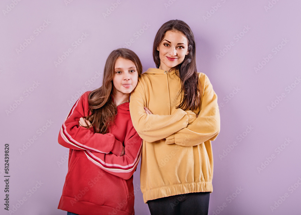Smiling fun mother and playful daughter showing the tongue. Front view with folded arms in fashion trendy hoodie together on purple studio background with empty copy space. Happy loving family Smiling fun mother and playful daughter showing the tongue. Front view with folded arms in fashion trendy hoodie together on purple studio background with empty copy space. Happy loving family