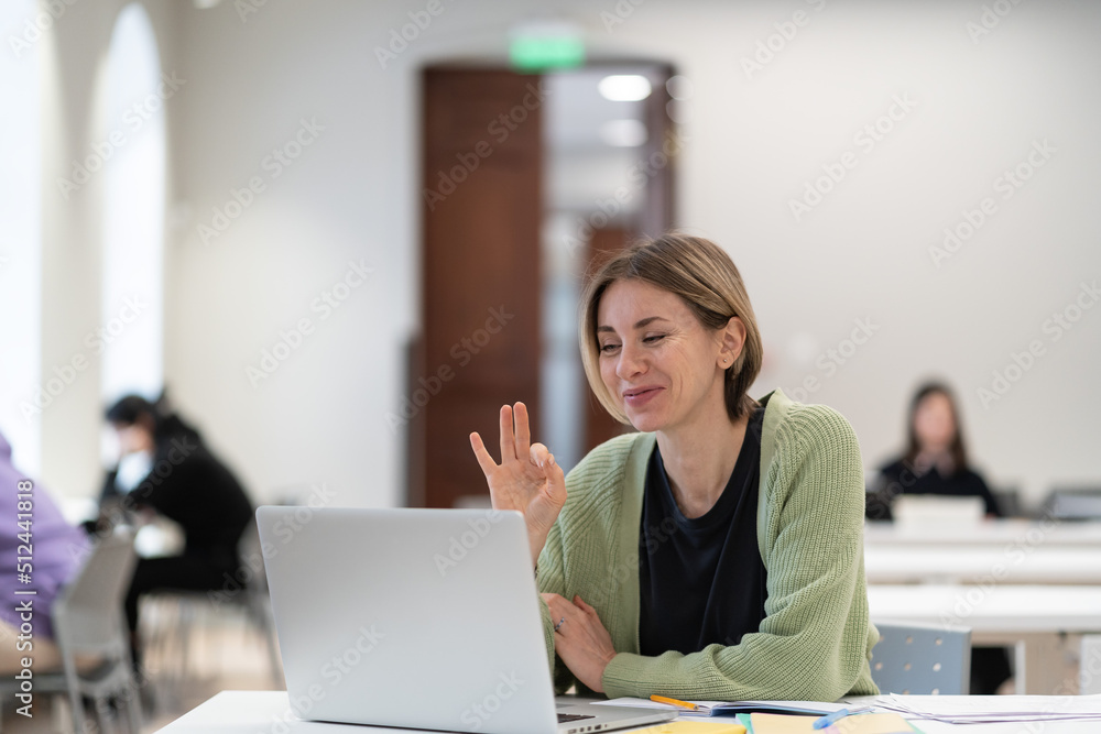 Happy female mature student showing ok gesture at web camera during virtual class, sitting at library table with laptop, middle-aged woman enjoying e-learning, getting second degree remotely Happy female mature student showing ok gesture at web camera during virtual class, sitting at library table with laptop, middle-aged woman enjoying e-learning, getting second degree remotely