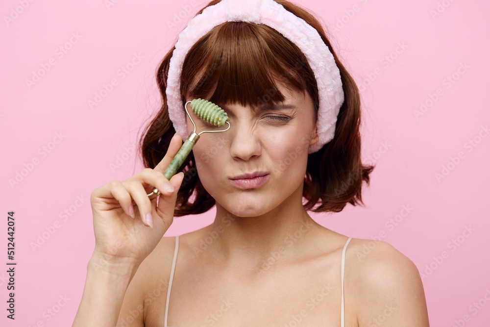An emotional woman with clean skin, an ideal smile and dark beautiful hair stands on a pink background with a beautiful bandage on her head with a facial massage roller.Horizontal studio shot. An emotional woman with clean skin, an ideal smile and dark beautiful hair stands on a pink background with a beautiful bandage on her head with a facial massage roller.Horizontal studio shot.