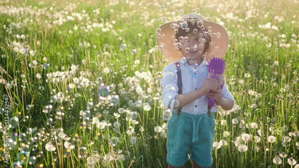 portrait of a five-year-old boy in a hat, stands on a field of dandelions and shoots soap bubbles from a toy gun at sunset in summer portrait of a five-year-old boy in a hat, stands on a field of dandelions and shoots soap bubbles from a toy gun at sunset in summer