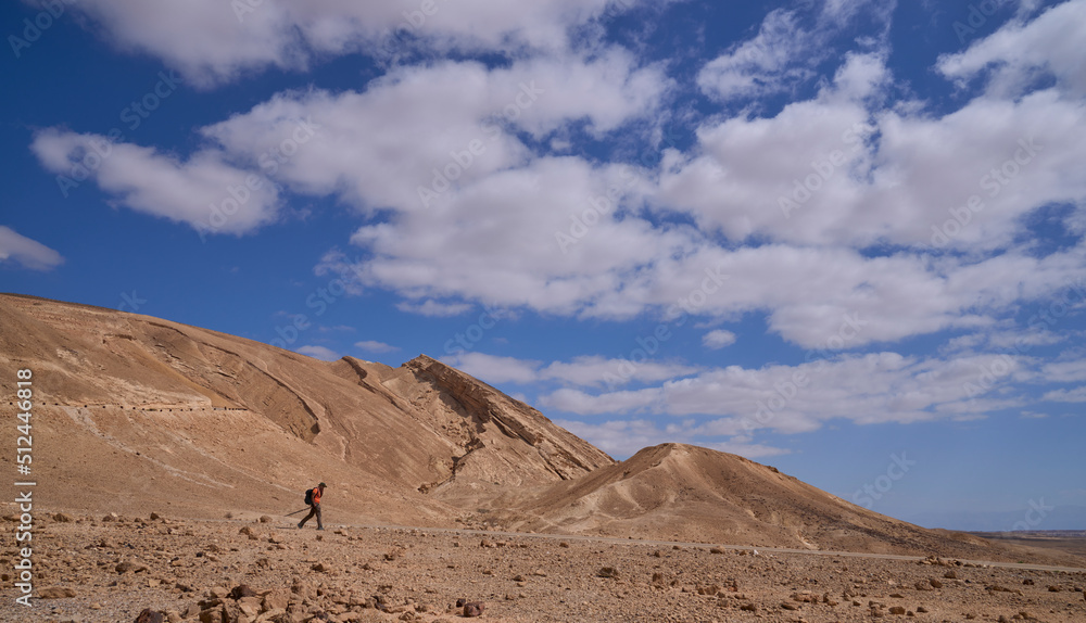 Male hiker on a trail in Negev desert. Orange and yellow colors of the mountains rocky landscape. Almost vertical wall of the Small Crater at the background. Popular hiking in park Hamakhtesh Hakatan.
