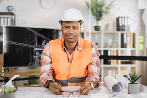 Attractive African engineer in white helmet and reflective vest sitting at table with lots of blueprints on the background of large tv screen with sketch. Business man working on construction project.