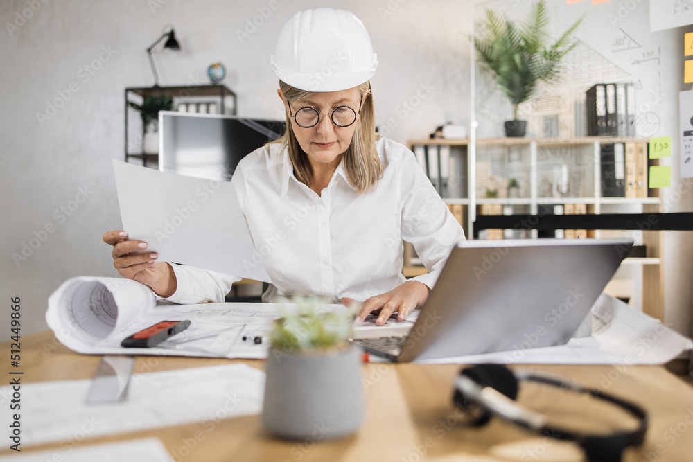 Senior woman builder, engineer in white shirt and hard hat on desk in office on background of large TV screen with, working on laptop while looking on papers or sketches. Senior woman builder, engineer in white shirt and hard hat on desk in office on background of large TV screen with, working on laptop while looking on papers or sketches.