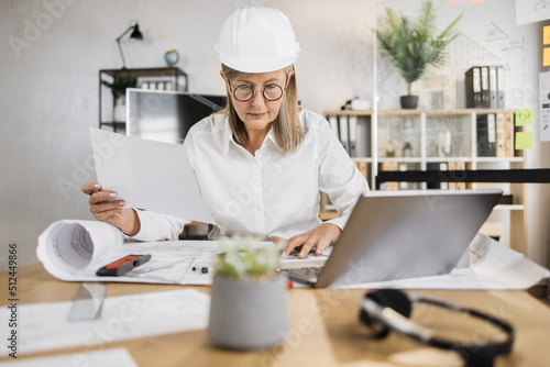Senior woman builder, engineer in white shirt and hard hat on desk in office on background of large TV screen with, working on laptop while looking on papers or sketches.