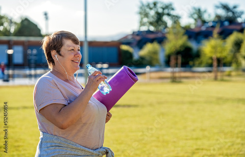 Happy smiling senior plus size woman with earphones holding yoga mat outdoors drinking water resting after exercises at warm sunny summer day
