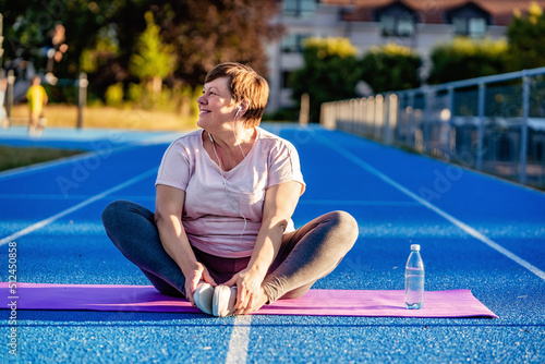 Relaxed senior plus size woman with earphones sitting on yoga mat on sports ground outdoors resting after exercises at warm sunny summer day