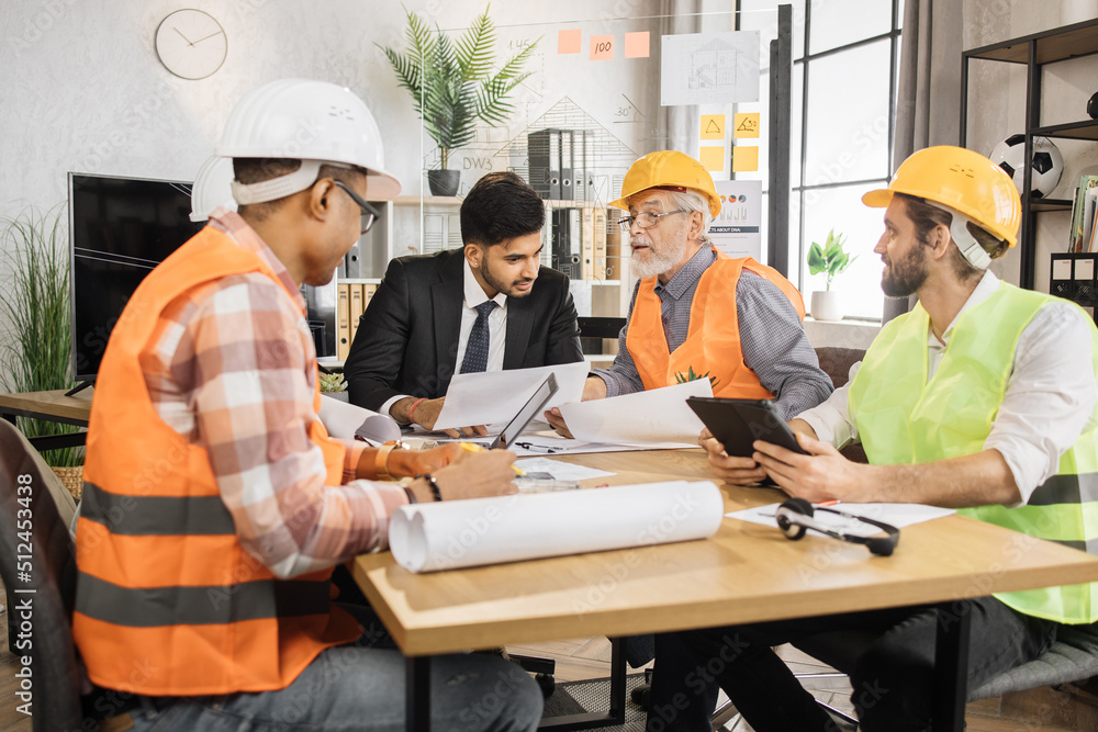 Multiracial men builders in formal outfit and helmets sitting at table with lots of blueprints during important strategic session. Business people working on common construction project. Multiracial men builders in formal outfit and helmets sitting at table with lots of blueprints during important strategic session. Business people working on common construction project.