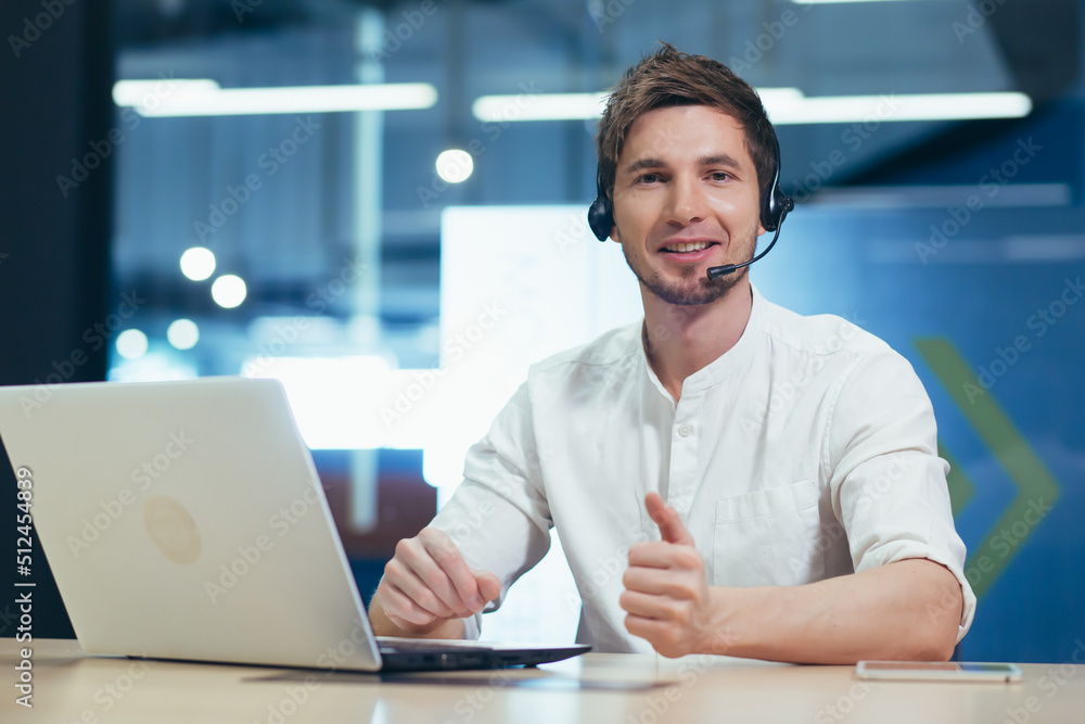 Video conference. A young handsome man in headphones sits in the office behind a laptop and communicates through a video call. He looks at the camera, smiles Video conference. A young handsome man in headphones sits in the office behind a laptop and communicates through a video call. He looks at the camera, smiles