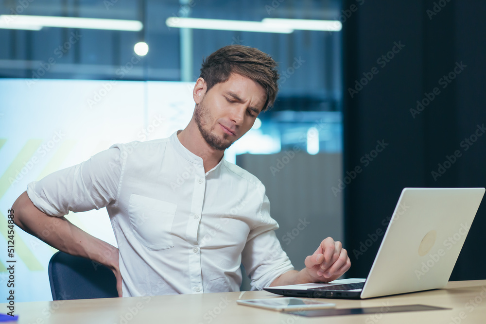 Feelings of fatigue in the workplace. Young man office worker, freelancer, IT specialist tired, holding his back, feeling pain, discomfort. Needs rest. Feelings of fatigue in the workplace. Young man office worker, freelancer, IT specialist tired, holding his back, feeling pain, discomfort. Needs rest.