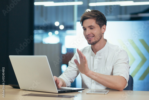 Working video call in the office. Young male businessman, freelancer sitting at a desk in the office on a laptop, talking through the camera on a video call, explaining something, waving his hands.