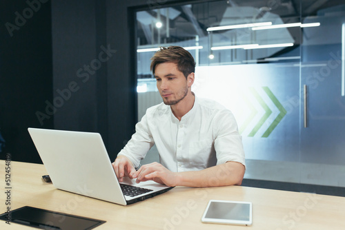 Working environment. Portrait of a young handsome male businessman. Sitting at a desk in the office, working on a laptop, tablet, cell phone. Gains, works.