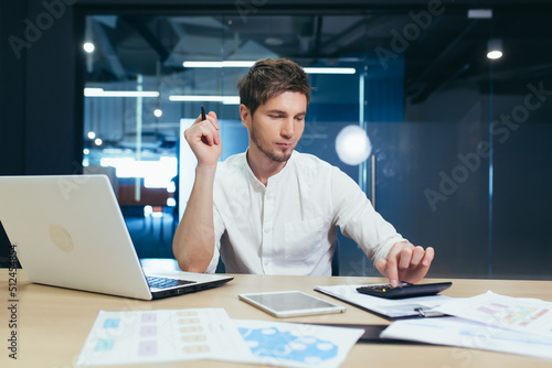 Young male accountant, economist, financier working in the office at the table, typing and counting on a calculator. Works with documents and laptop. Satisfied with the work