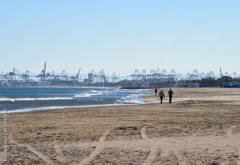Сoastline against backdrop of gantry cranes and shipping containers in port. Waves in sea at coast. beach near seaport. People walking beach at sea. Seashore with storm waves in winter at resort. .