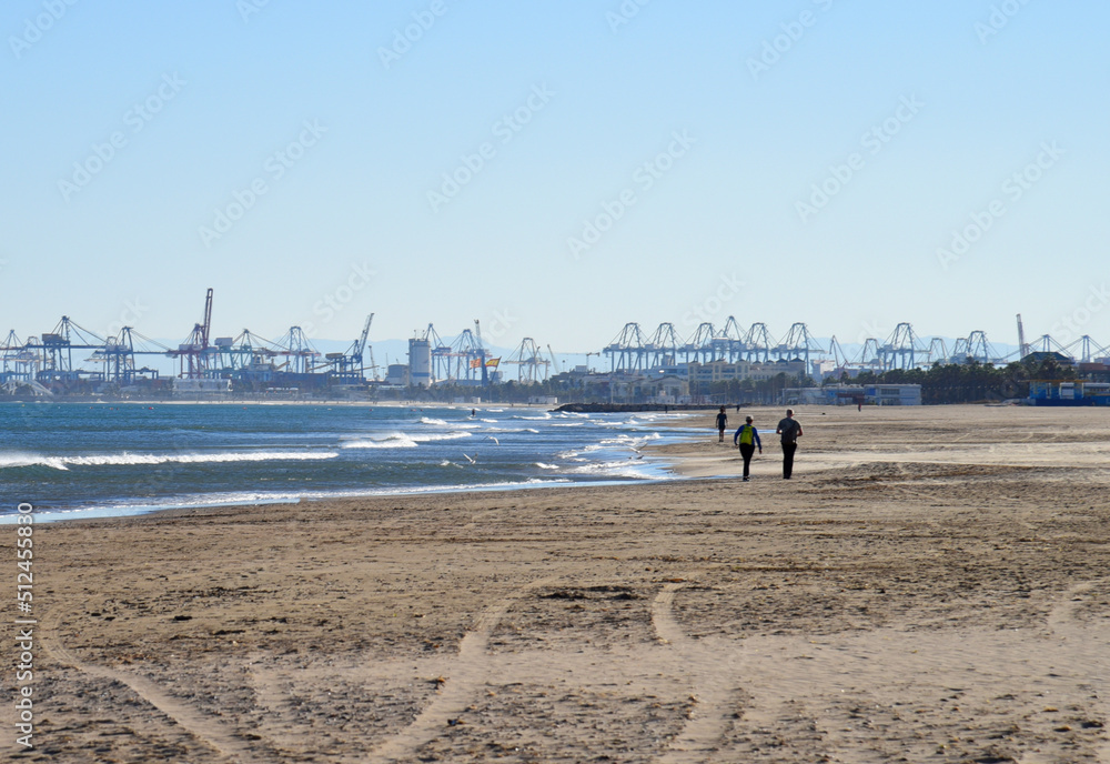 Сoastline against backdrop of gantry cranes and shipping containers in port. Waves in sea at coast. beach near seaport. People walking beach at sea. Seashore with storm waves in winter at resort. . Сoastline against backdrop of gantry cranes and shipping containers in port. Waves in sea at coast. beach near seaport. People walking beach at sea. Seashore with storm waves in winter at resort. .