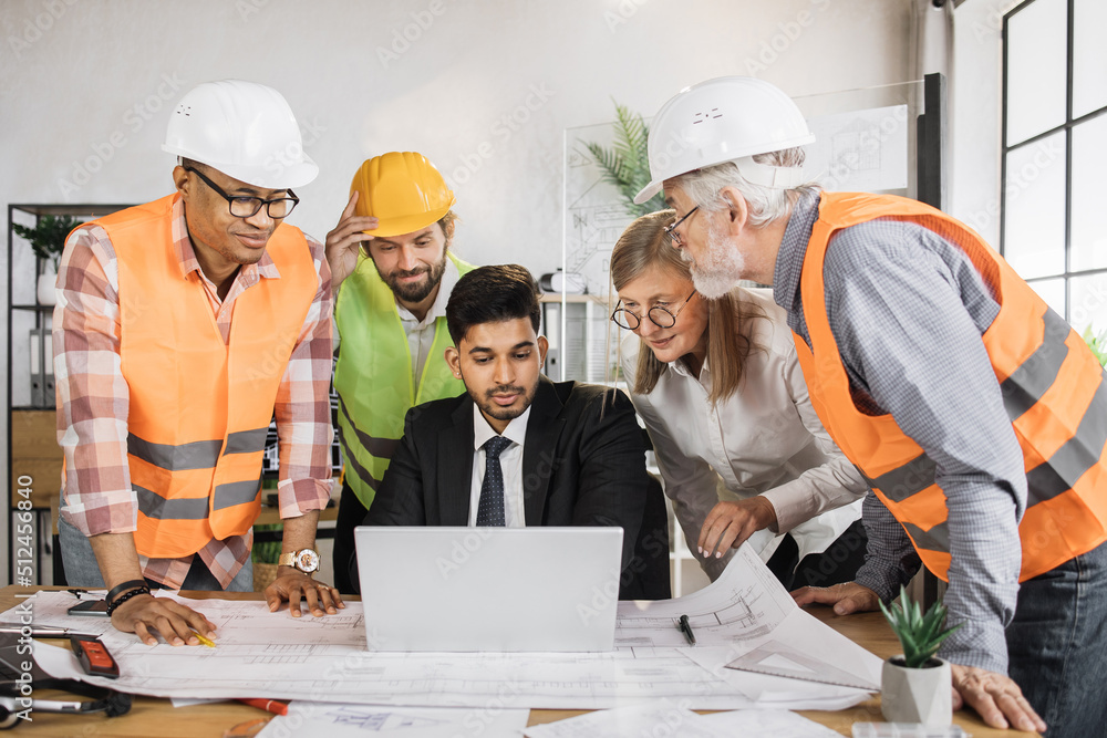 Four men and one woman sitting together at table and discussing blueprints on laptop during business conference. Multi ethnic architects, designers and engineers working at office. Four men and one woman sitting together at table and discussing blueprints on laptop during business conference. Multi ethnic architects, designers and engineers working at office.