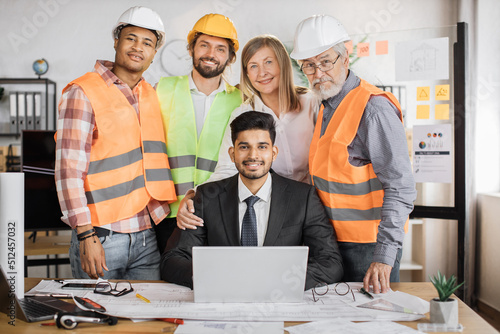 Team of qualified architects, engineers and designers having meeting in boardroom at office indoor, standing near indian man boss, sitting on desk with laptop, looking at camera. © sofiko14