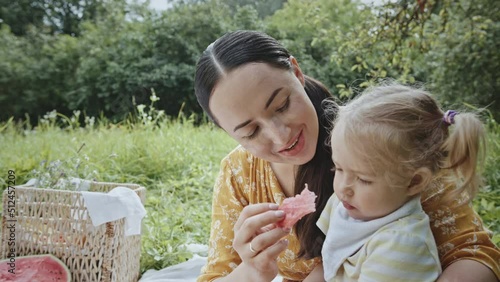 Mother feeding daughter during picnic. Adult mom with dark hair trying to give peach and watermelon slice to capricious little daughter while having picnic on summer weekend day in countryside