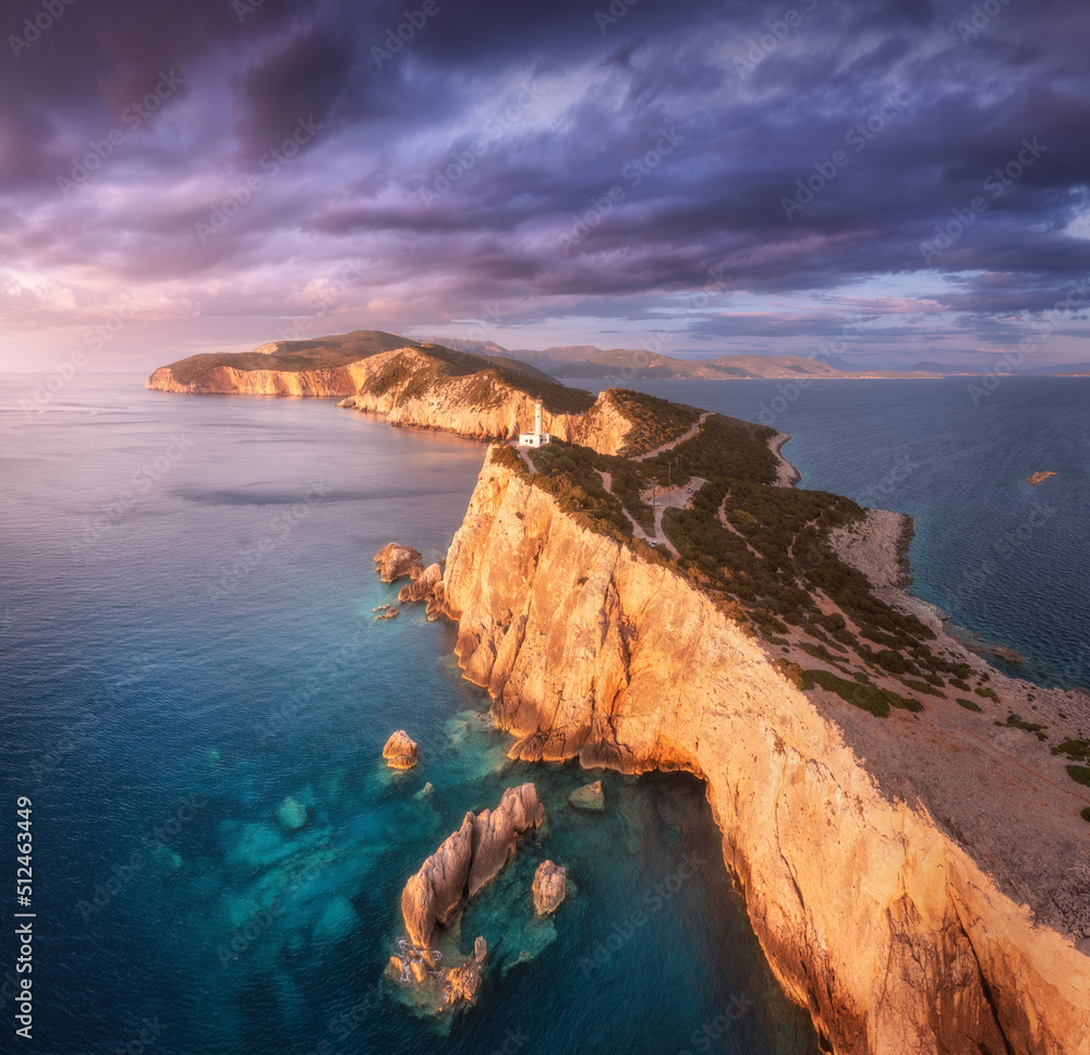 Lighthouse on the mountain peak at colorful sunset in summer. Aerial view. Beautiful lighthouse on the rock, sea and moody sky with purple clouds at dusk. Top view of Cape Lefkada, Greece. Landscape