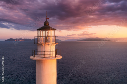 Lighthouse on the mountain peak at colorful sunset in summer. Aerial view. Beautiful lighthouse, sea and orange sky with pink and purple clouds at dusk. Top view of Cape Lefkada, Greece. Landscape