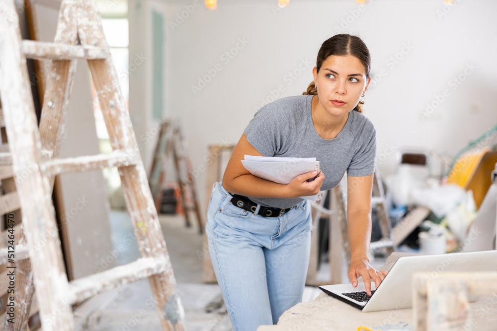 Young woman designer checking documents and using her laptop during repair works indoors.